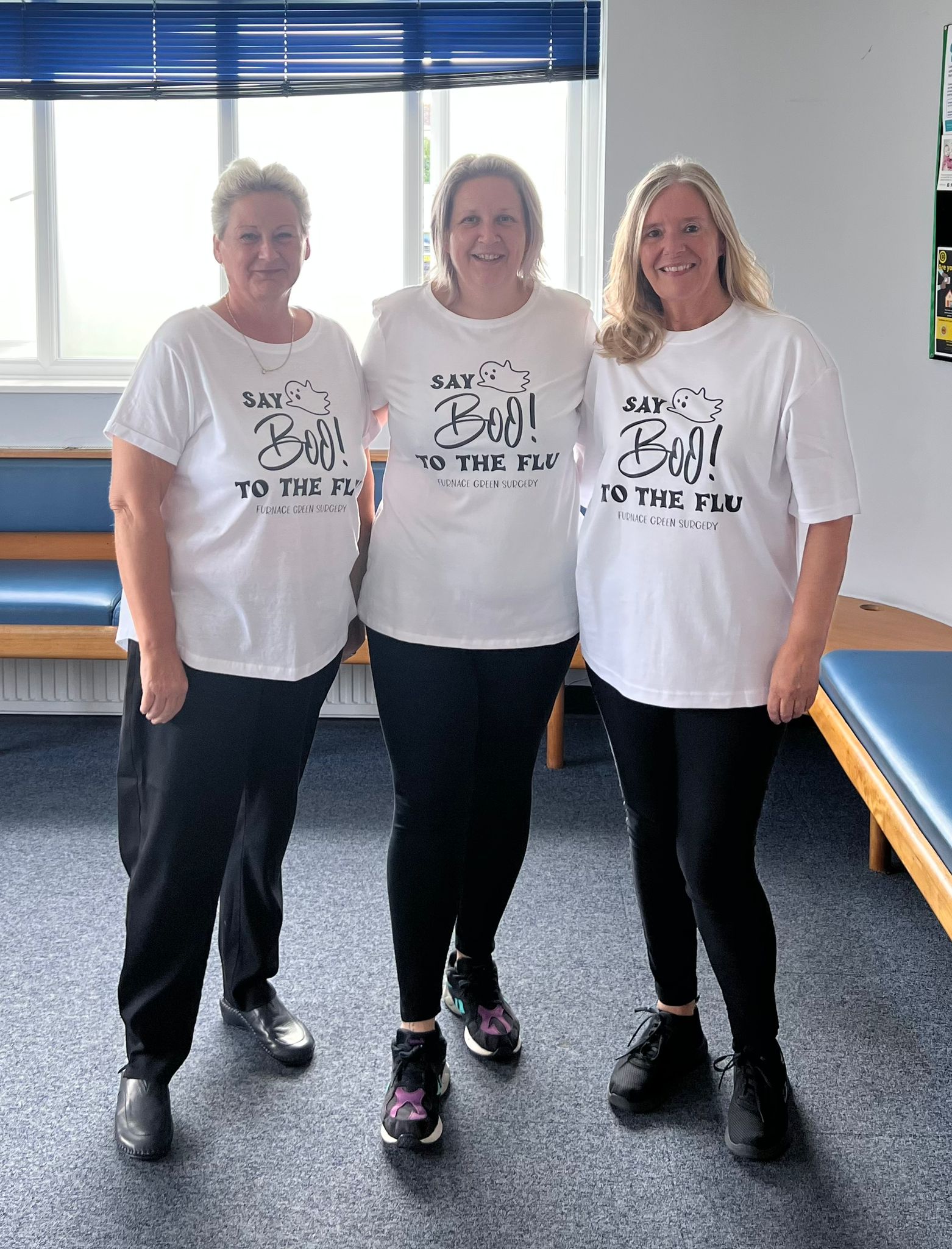 A photograph of three of our nurses in our reception. They are all wearing white t-shirts with a design printed in black. It says "Say Boo! to the flu" with a little cartoon ghost flying above the word 'Boo!' Below the design it says Furnace Green Surgery. They are all smiling at the camera.